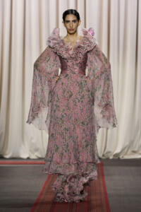 Model on a runway wearing a pink floral gown with large ruffled neckline and sheer cape sleeves.