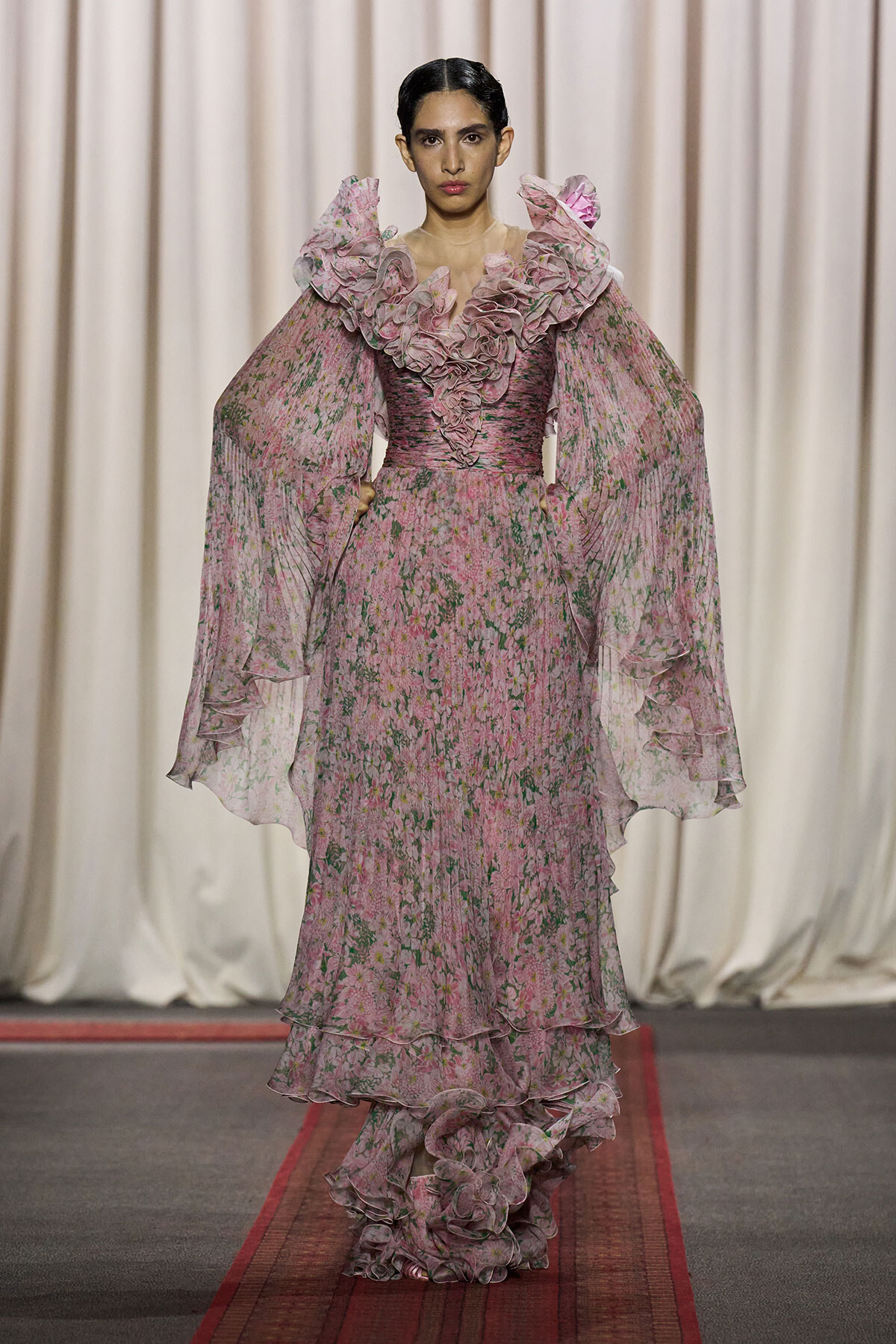 Model on a runway wearing a pink floral gown with large ruffled neckline and sheer cape sleeves.