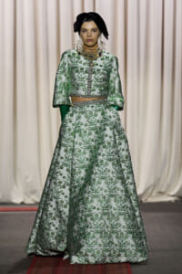 Model on a runway wearing a pale green, botanical-embroidered two-piece gown with a cropped top, full skirt, and matching belt, plus green gloves and a black head wrap.