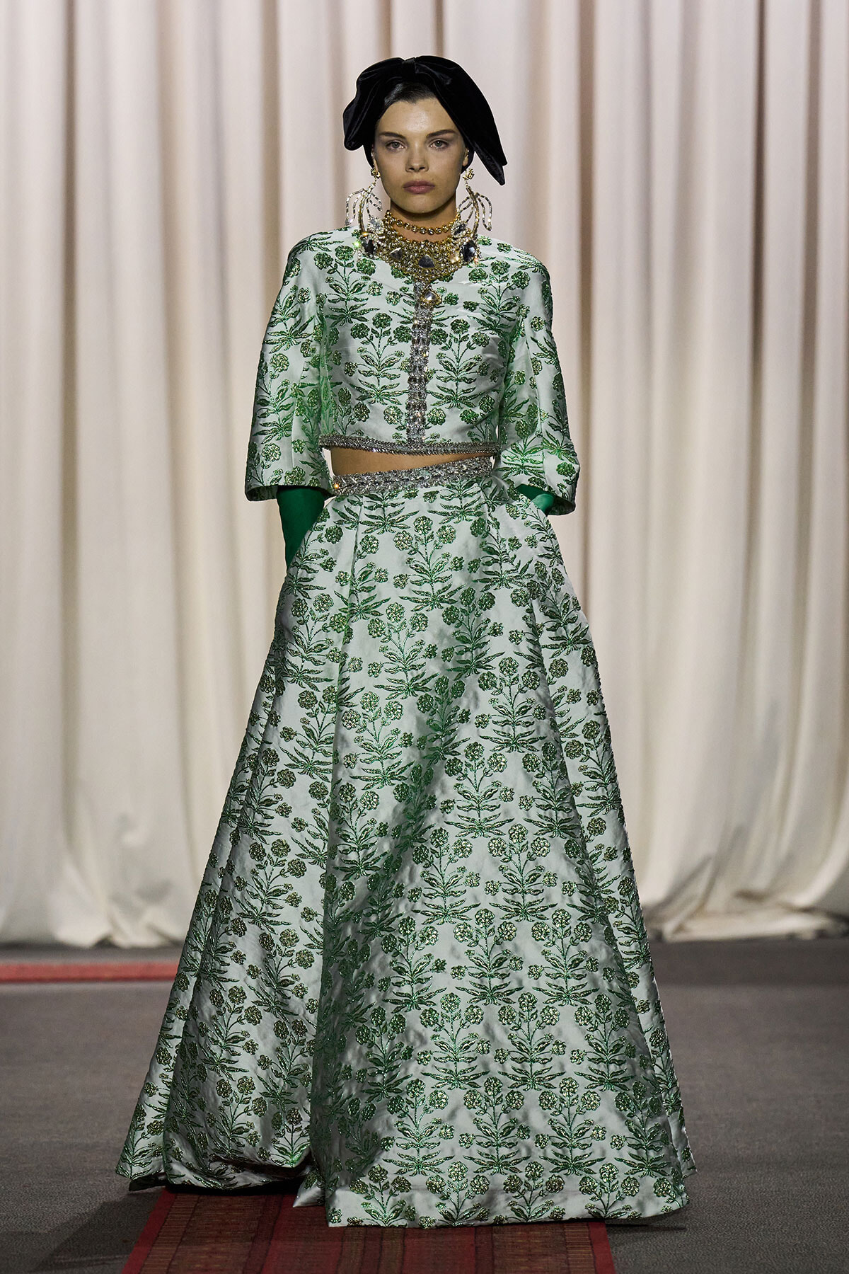 Model on a runway wearing a pale green, botanical-embroidered two-piece gown with a cropped top, full skirt, and matching belt, plus green gloves and a black head wrap.