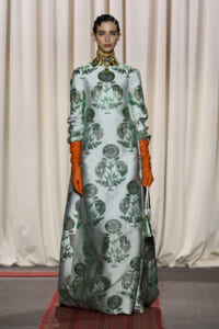 Model walking a runway in a pale blue, floor-length gown with green botanical embroidery, complemented by orange gloves and a chunky gold choker.