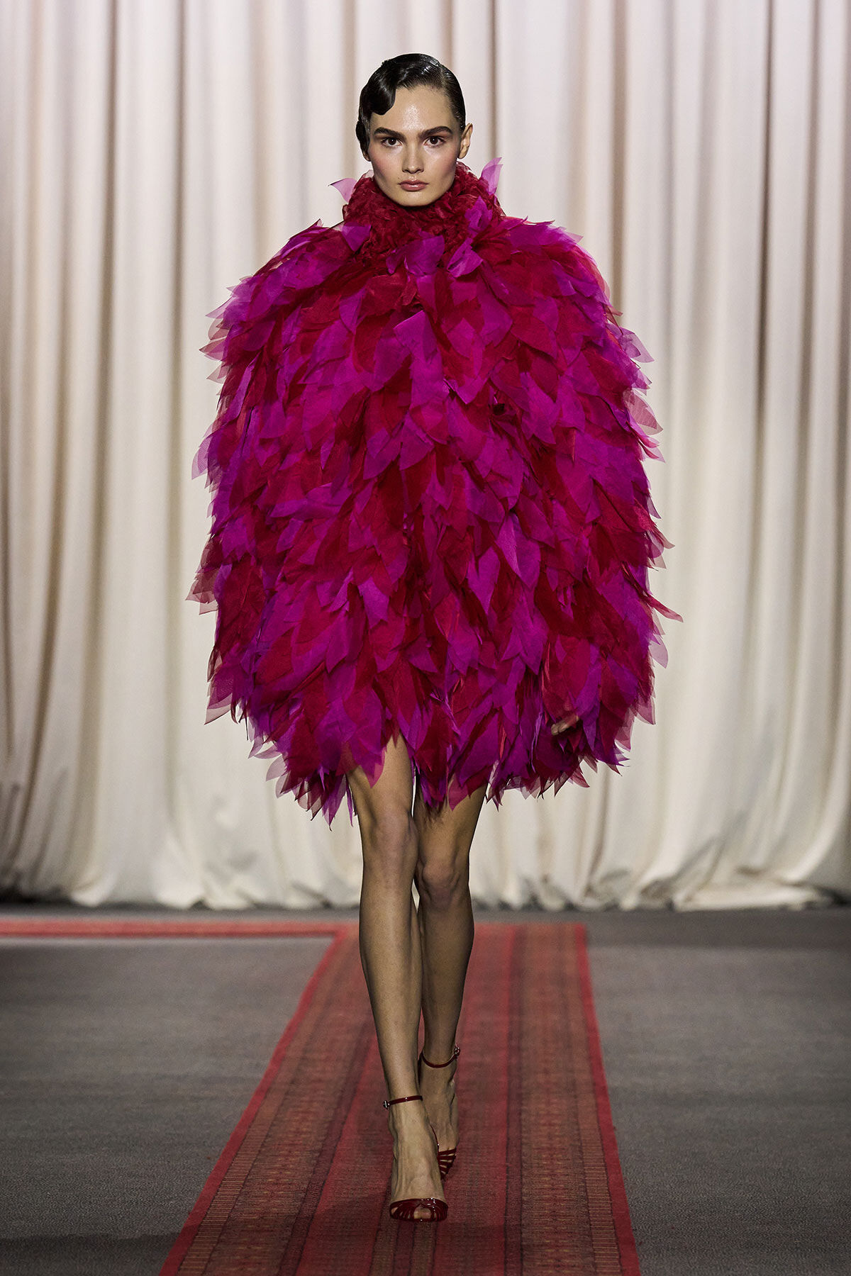 Model on runway wearing a vivid magenta feathered cape dress with a high neck.