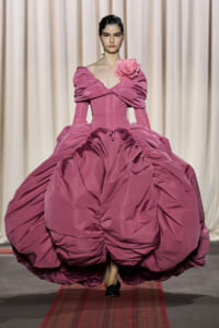Model walking a runway in a voluminous pink ball gown with a structured bodice and a pink flower at the shoulder