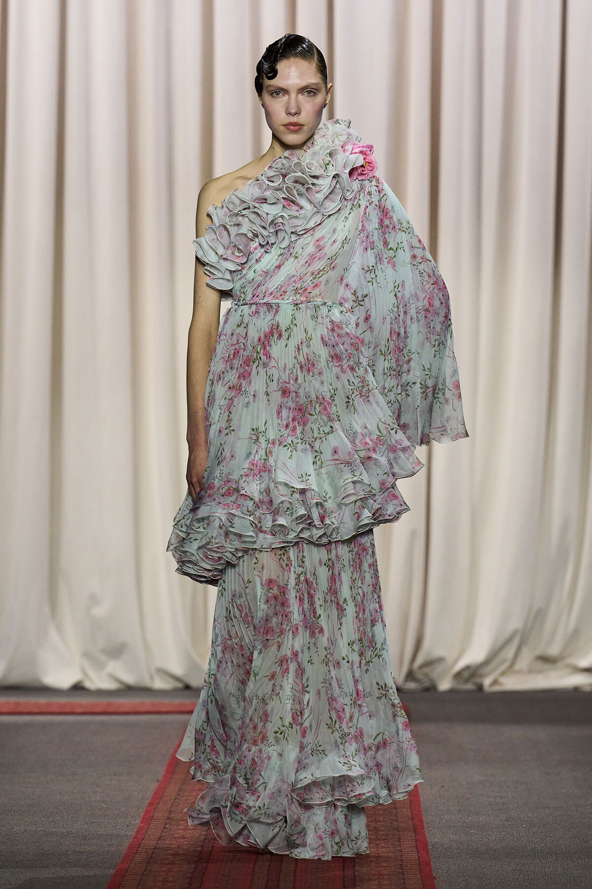 Model on a runway wearing a pale floral one-shoulder gown with cascading ruffles and a layered, tulle-like skirt.