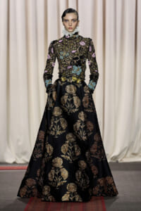 Model on a runway wearing a black gown with gold floral embroidery and a high ruffled collar.