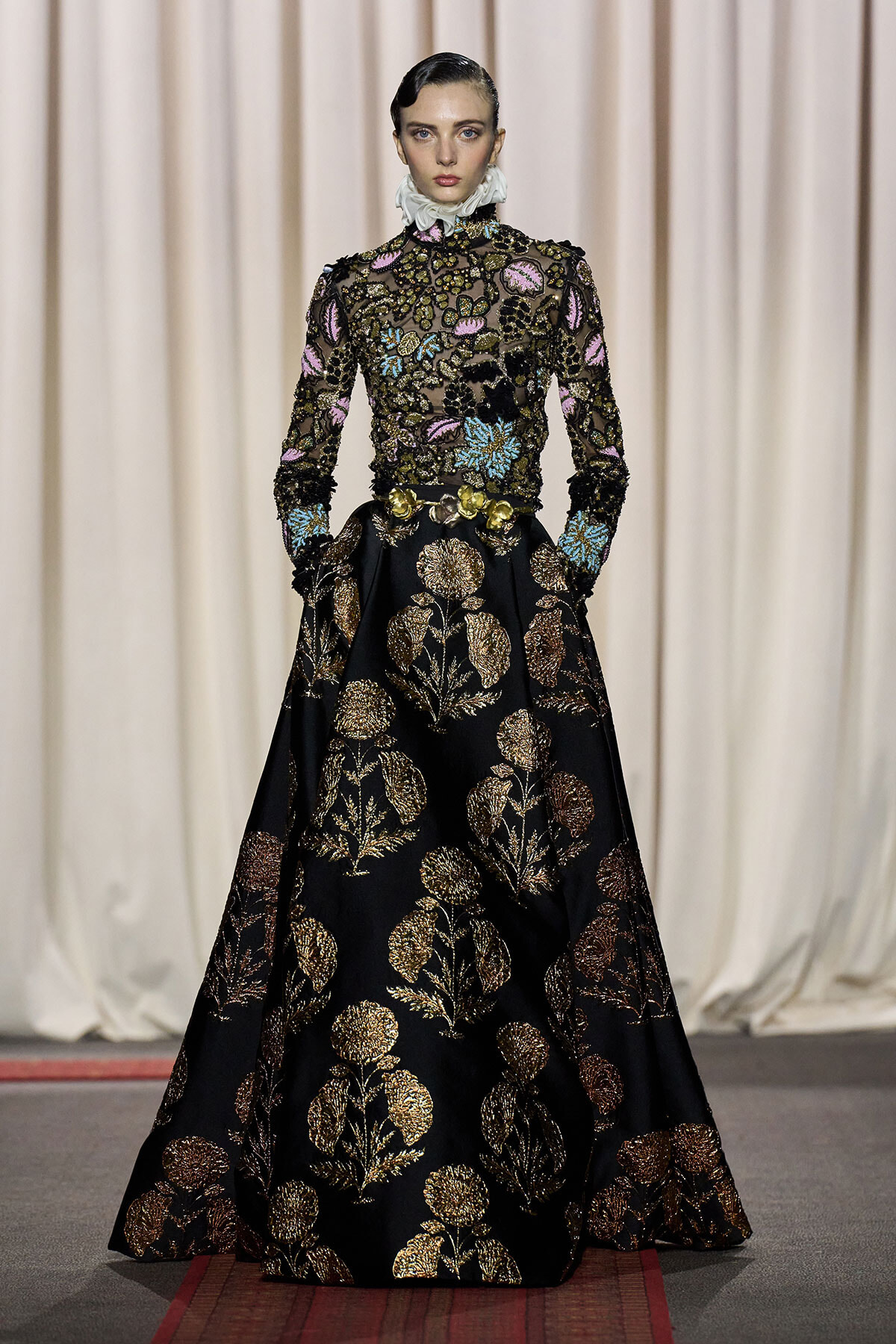 Model on a runway wearing a black gown with gold floral embroidery and a high ruffled collar.