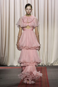 Model walking the runway in a pale pink, multi-tiered tulle gown with a ruffled capelet and waist cutout, paired with matching jewelry.