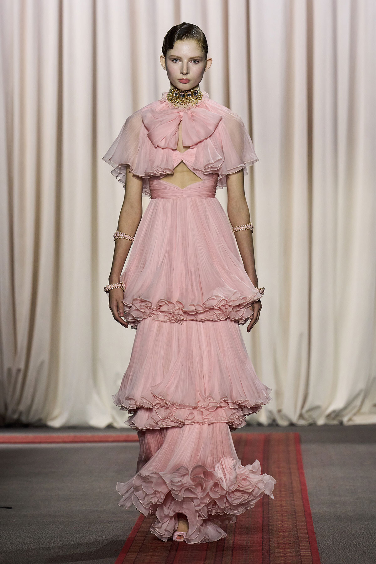 Model walking the runway in a pale pink, multi-tiered tulle gown with a ruffled capelet and waist cutout, paired with matching jewelry.