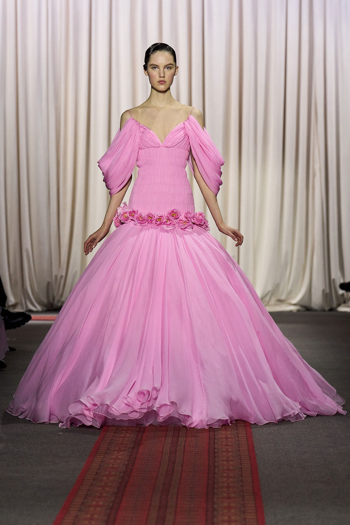 Model on a runway wearing a pink off-shoulder ball gown with voluminous sleeves and a floral belt, against a draped backdrop.