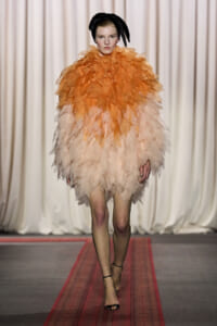 Model walking a runway in an orange-to-peach feathered dress with a black headwrap and strappy heels