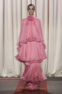 Model on a runway wearing a voluminous pink tulle gown with multiple ruffled layers and a rose-like flower at the high neck, against a neutral curtain backdrop.