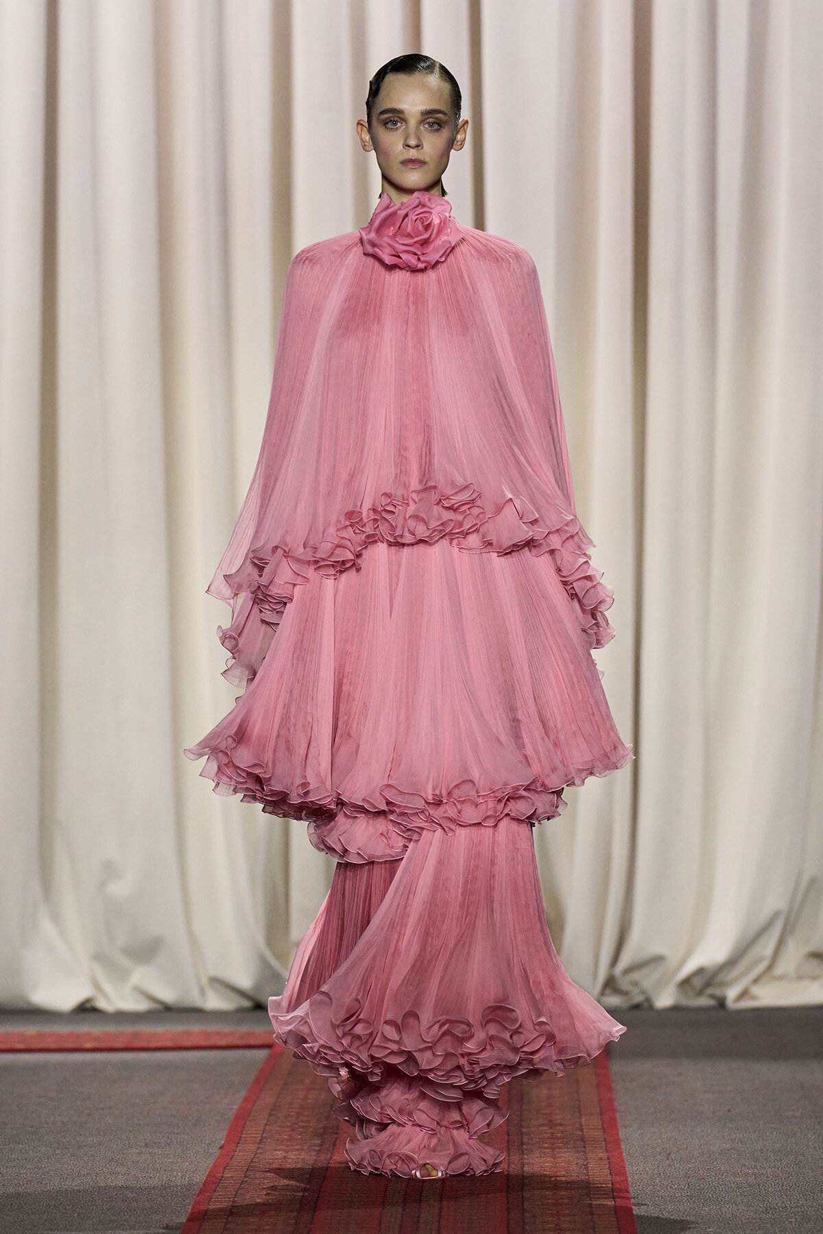 Model on a runway wearing a voluminous pink tulle gown with multiple ruffled layers and a rose-like flower at the high neck, against a neutral curtain backdrop.