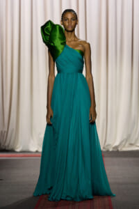 Model on a runway wearing a teal, full-length, one-shoulder gown with a dramatic emerald-green satin ruffle on the left shoulder.