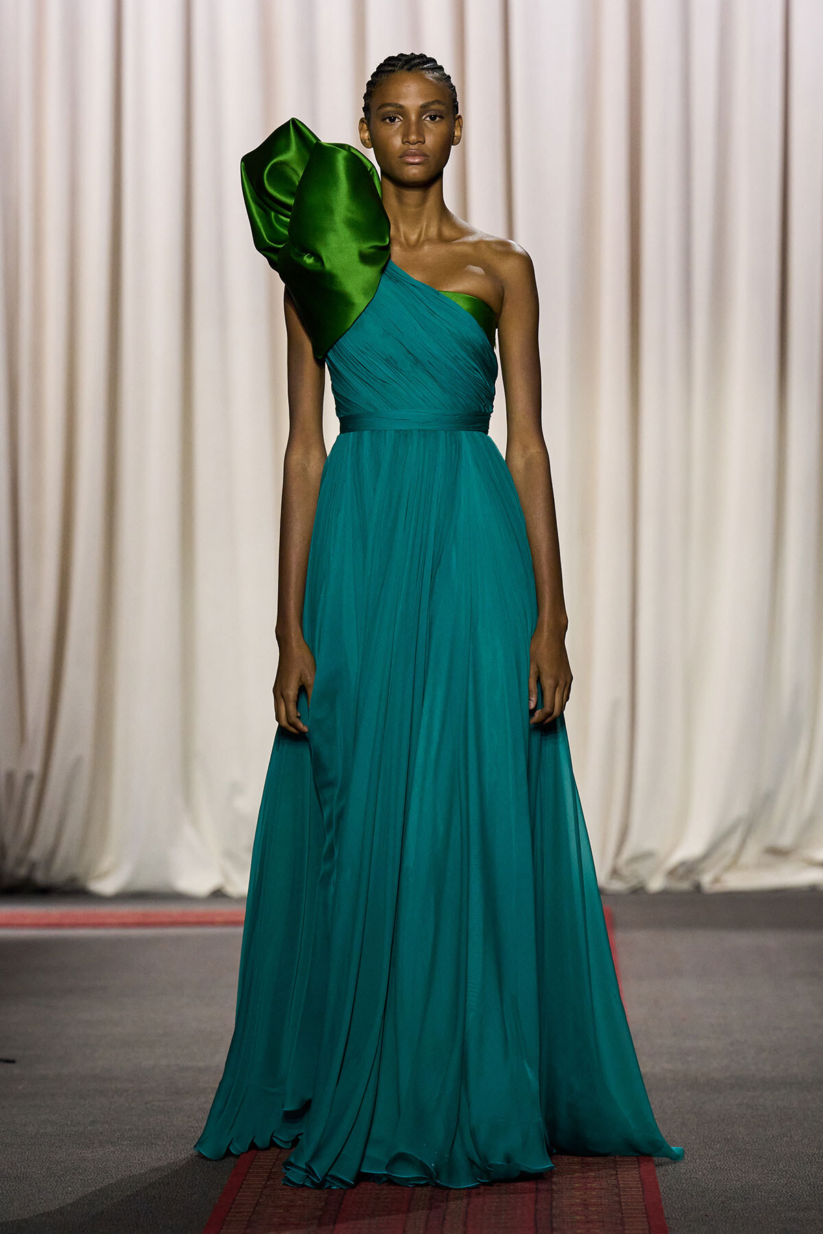 Model on a runway wearing a teal, full-length, one-shoulder gown with a dramatic emerald-green satin ruffle on the left shoulder.