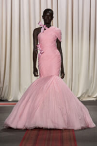 Dark-skinned model on a runway wearing a pale pink, one-shoulder ruched mermaid gown with bows at shoulder and waist.