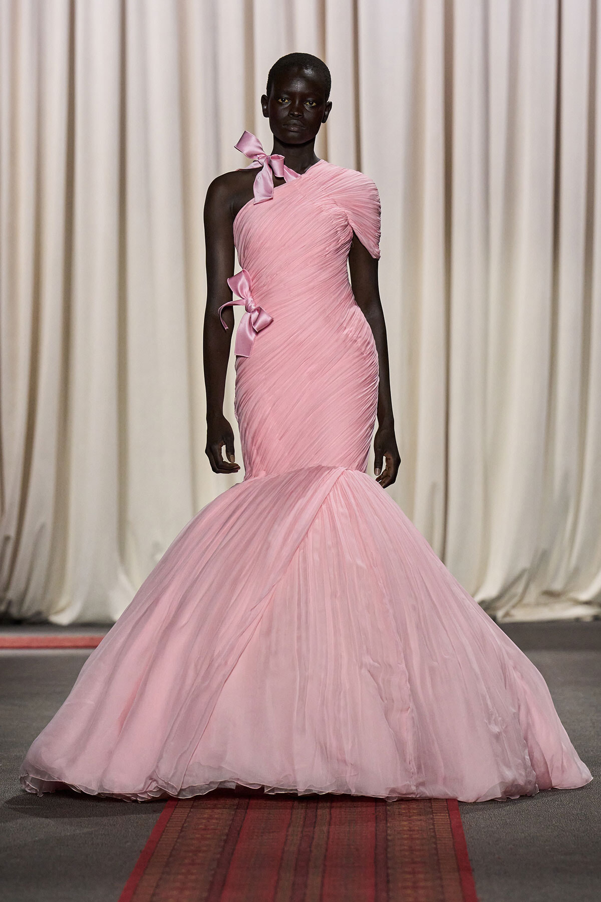 Dark-skinned model on a runway wearing a pale pink, one-shoulder ruched mermaid gown with bows at shoulder and waist.