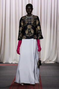 Model on runway wearing a black and gold embroidered top, a pale blue floor-length skirt, bright pink gloves, and a small handbag.