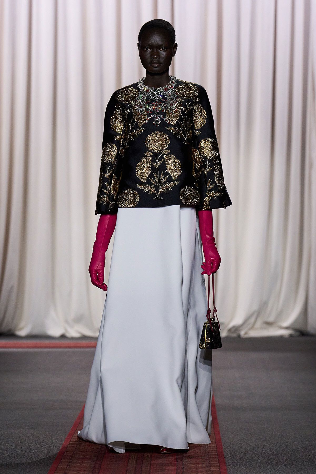Model on runway wearing a black and gold embroidered top, a pale blue floor-length skirt, bright pink gloves, and a small handbag.