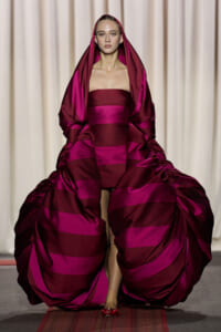 Model on runway wearing a bold magenta and burgundy striped gown with a voluminous robe draped over her shoulders and hooded headdress.