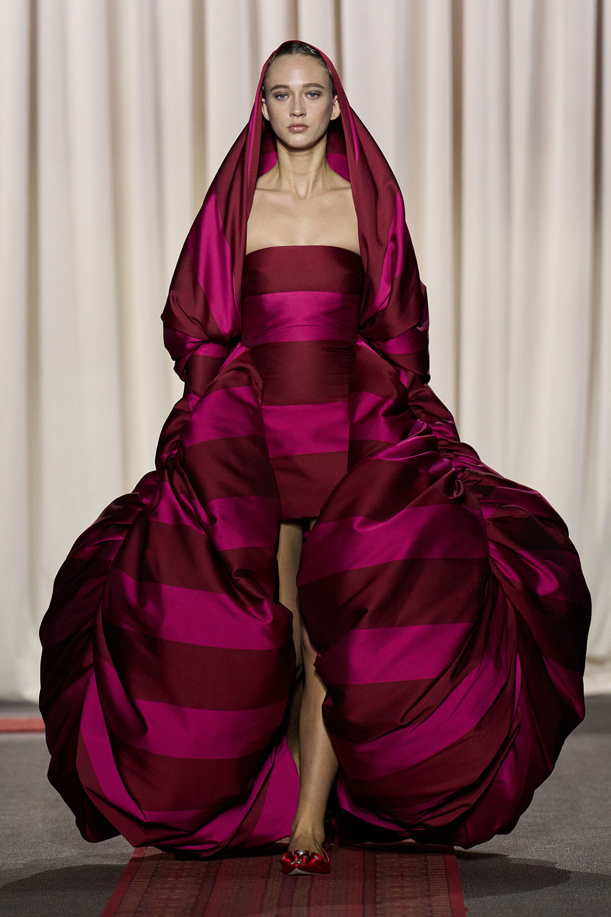 Model on runway wearing a bold magenta and burgundy striped gown with a voluminous robe draped over her shoulders and hooded headdress.