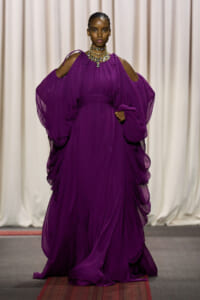 Model walks a fashion show wearing a flowing royal purple gown with dramatic draped sleeves and a statement necklace.