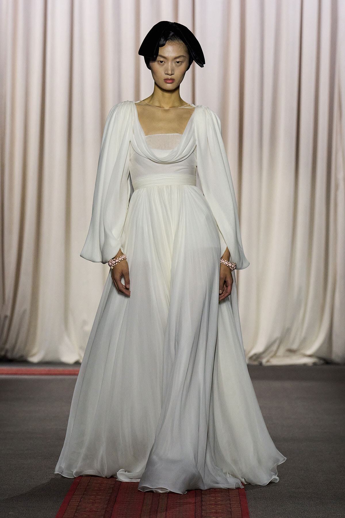 Model wearing a white, floor-length draped gown with voluminous sleeves on a fashion runway, accessorized with a black headpiece.