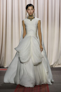 Model on a runway wearing a white ruched bodice gown with voluminous draped layers and a bold multi‑strand necklace.