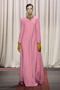 Model walking a fashion runway in a floor-length pink gown with wide sleeves, paired with a large jeweled necklace and yellow gloves.