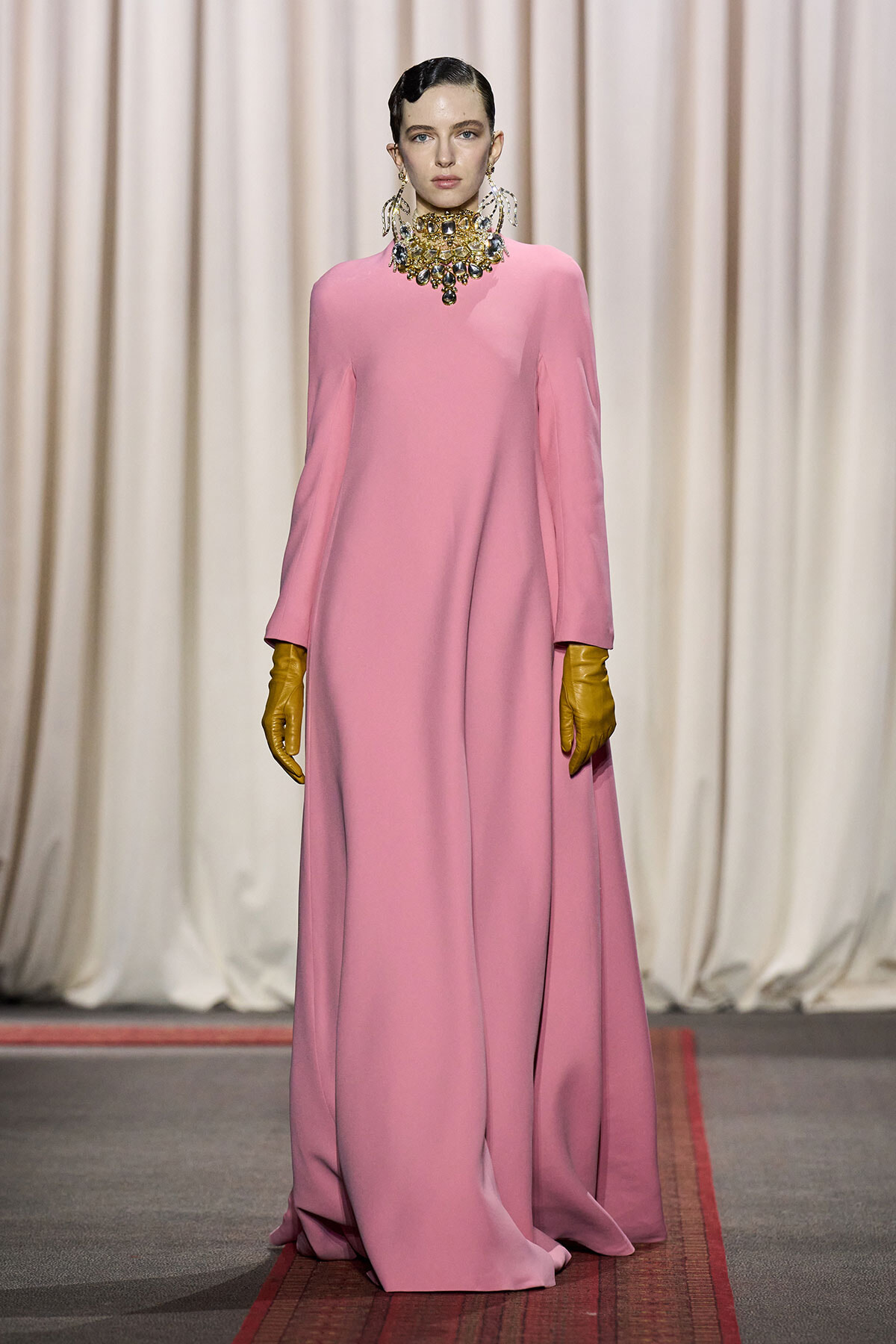 Model walking a fashion runway in a floor-length pink gown with wide sleeves, paired with a large jeweled necklace and yellow gloves.