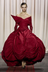 Model walking a runway in a dramatic burgundy ball gown with an off-the-shoulder neckline and voluminous, ruched skirt.