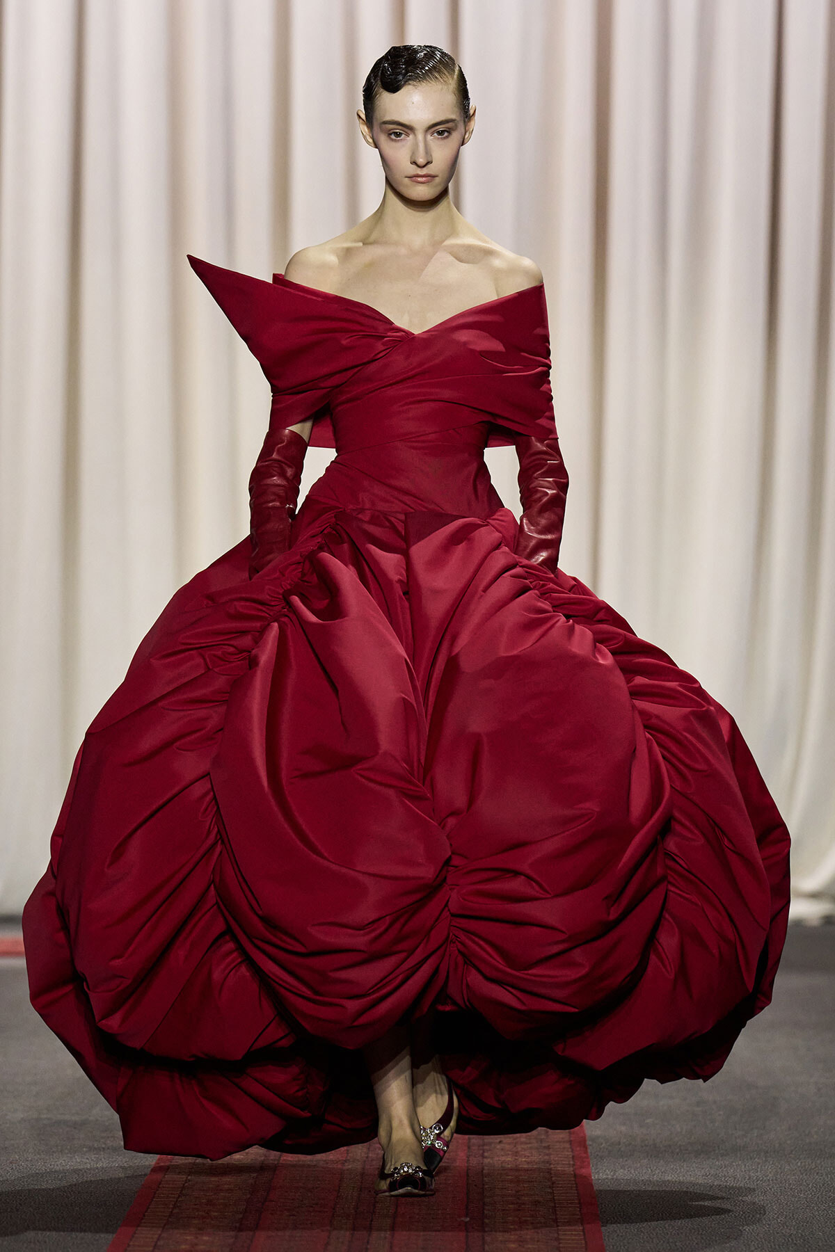 Model walking a runway in a dramatic burgundy ball gown with an off-the-shoulder neckline and voluminous, ruched skirt.