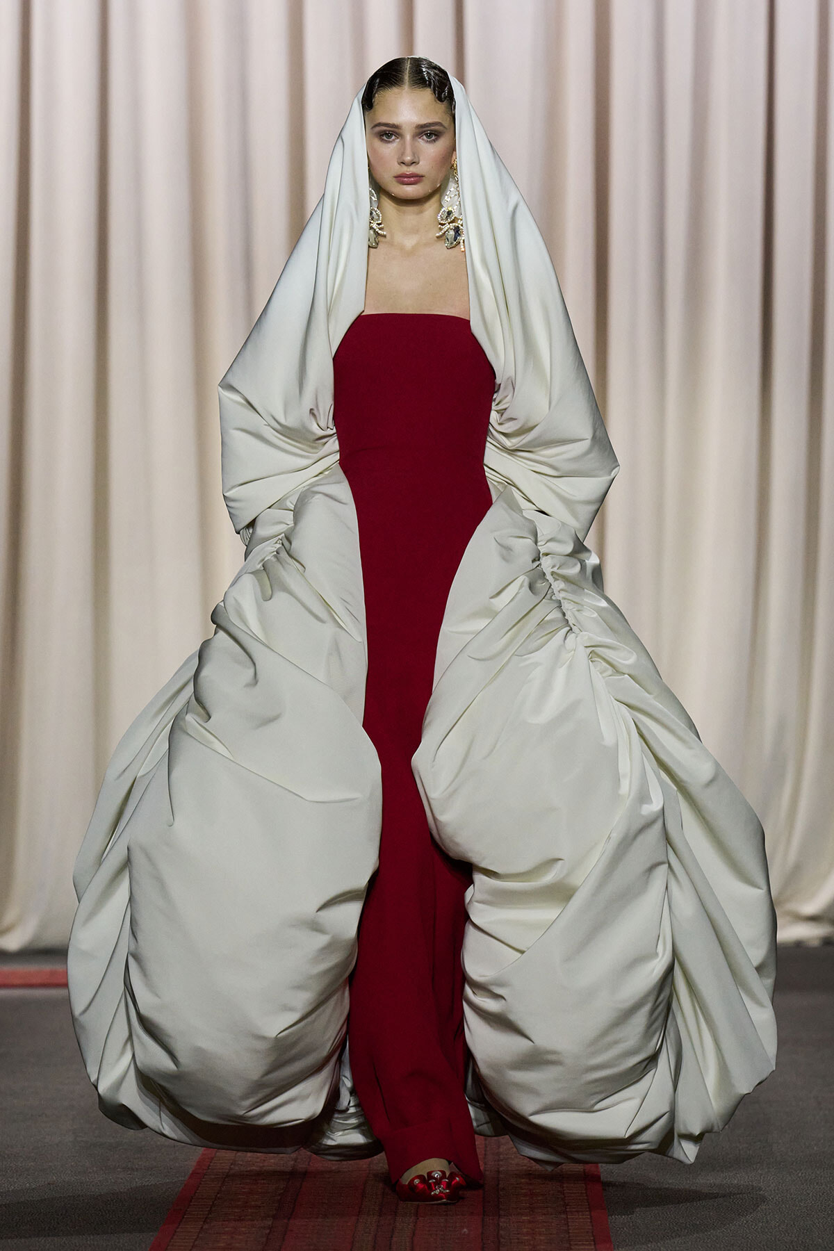 Model walks the runway in a strapless red gown with large ivory sculptural cape and statement earrings; dramatic fashion moment.