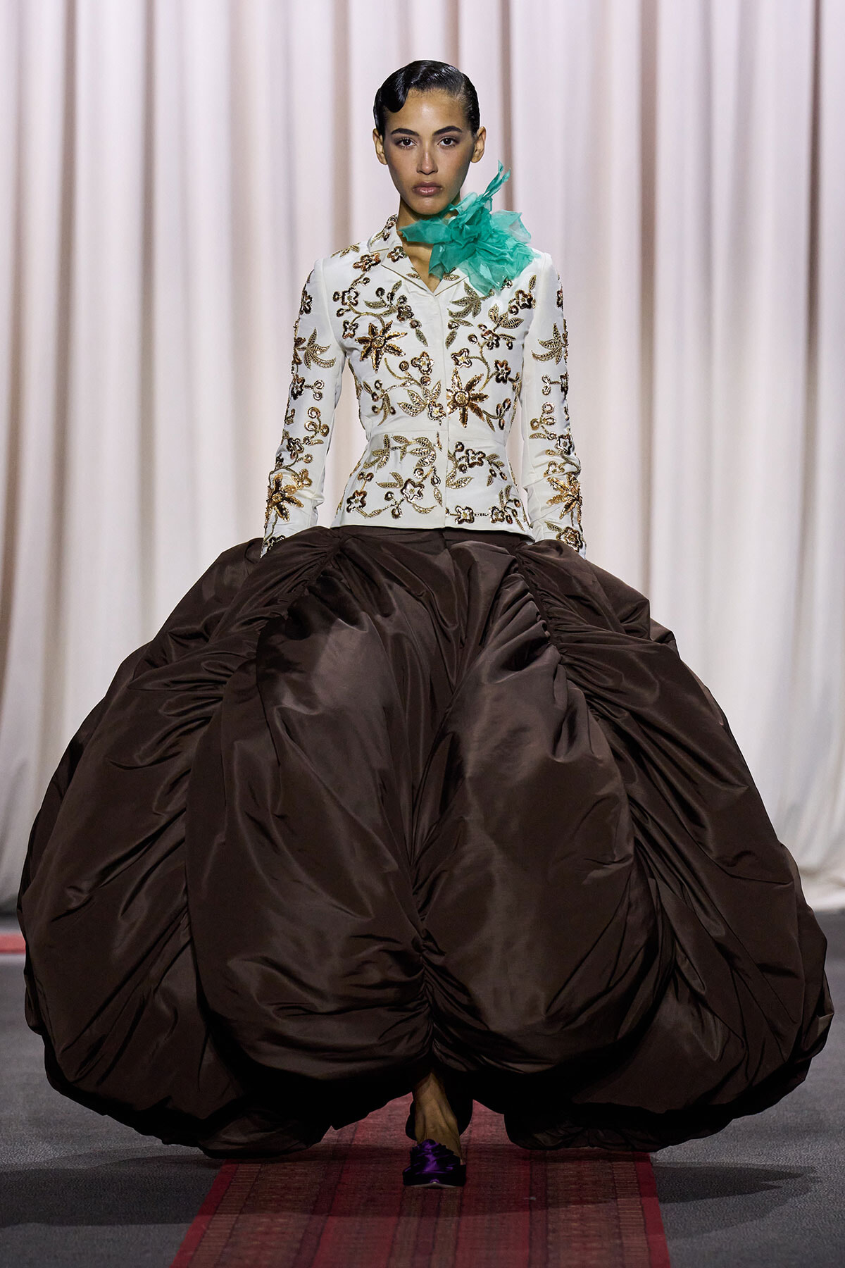 Model walking the runway in a white embroidered jacket with gold floral patterns and a dramatic brown ball gown skirt, teal neckpiece at the side.