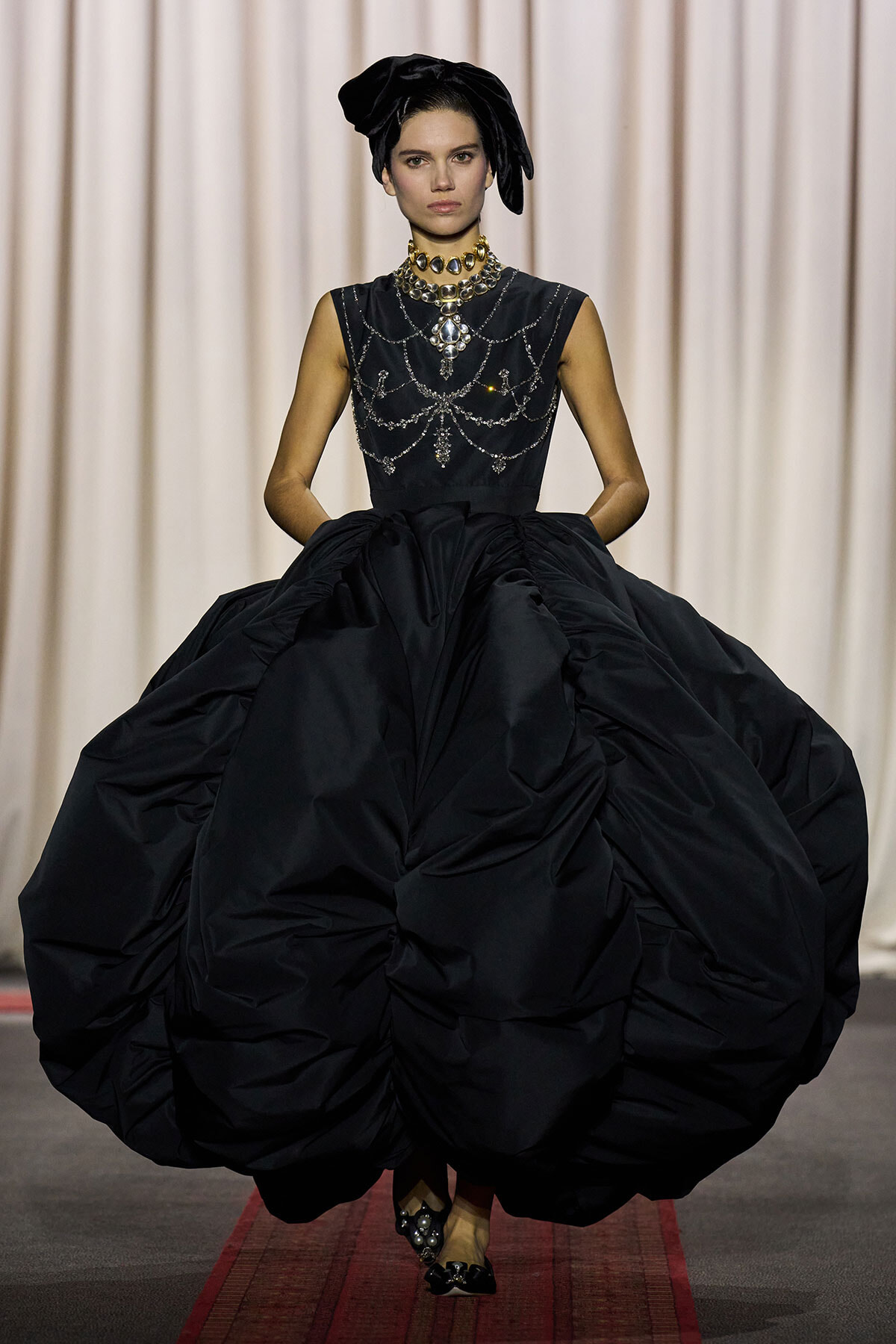 Runway model wearing a dramatic black satin ball gown with a jeweled bodice, gold necklaces, and a black head wrap on a catwalk with a beige curtain backdrop.