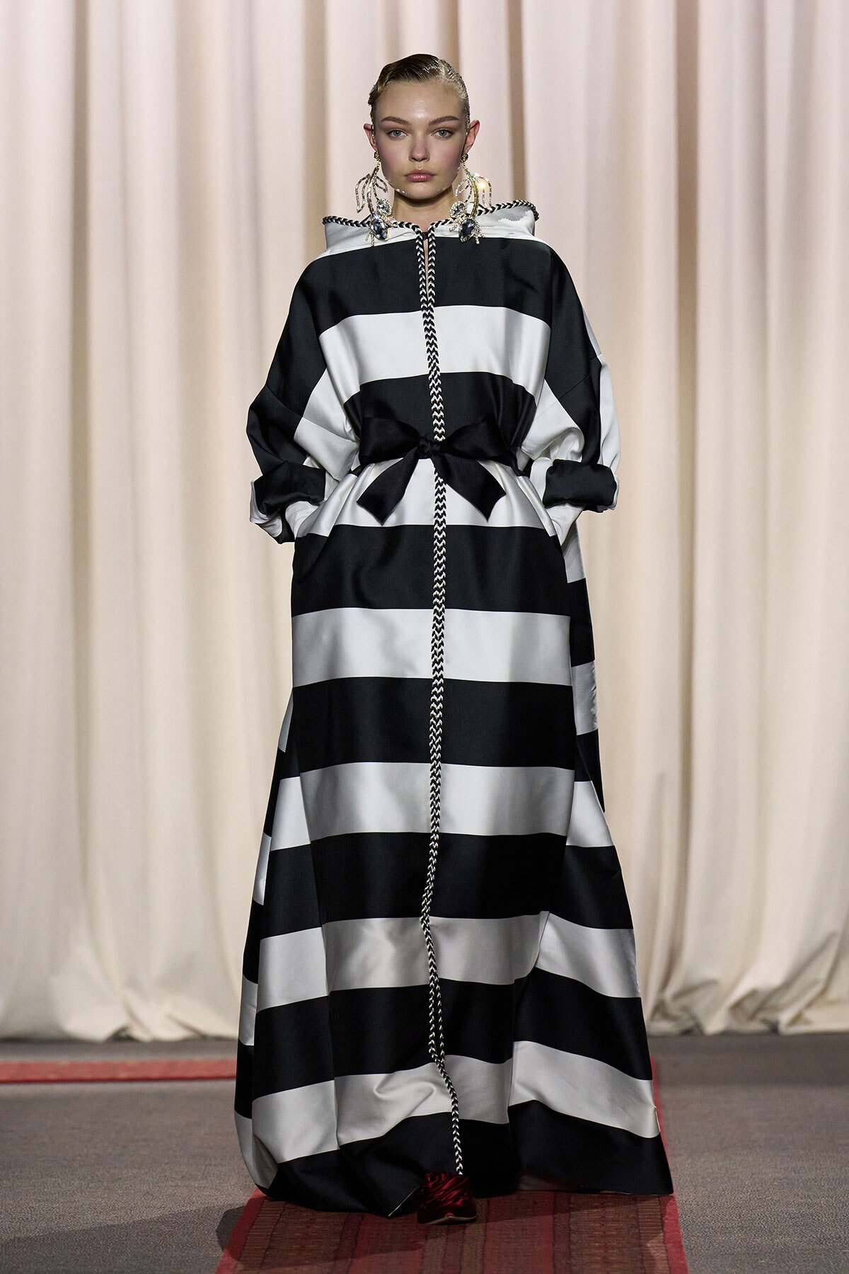 Runway model wearing a black-and-white striped, floor-length dress with a braided rope trim and a waist bow.