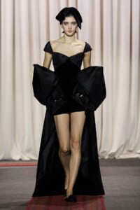 Model on a runway wearing a black off-shoulder gown with oversized sculptural sleeves and a long train.