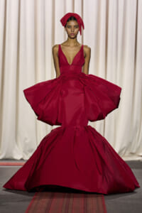 Model on a runway wearing a deep red ball gown with a plunging neckline and a voluminous, sculpted skirt.
