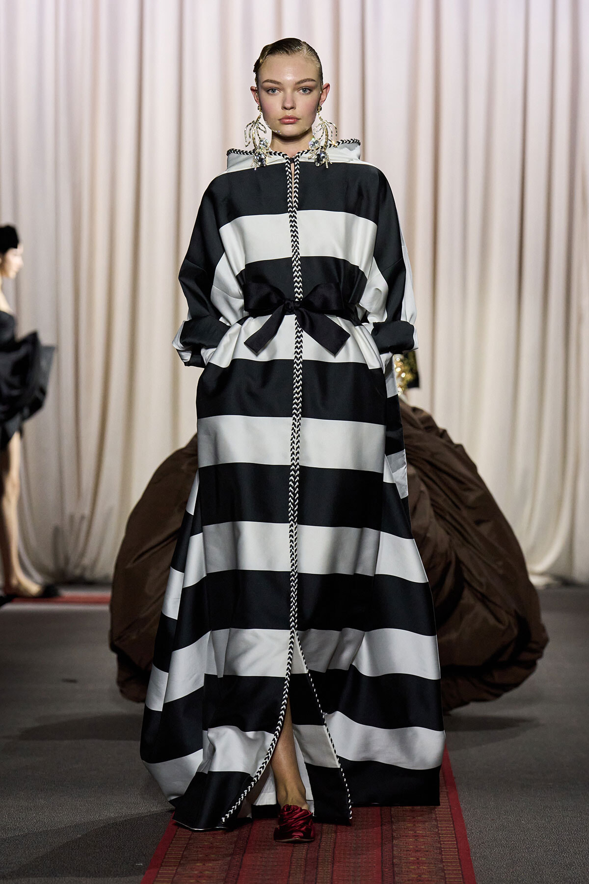 Runway model in a black-and-white striped maxi dress with a waist bow and braided front cord.