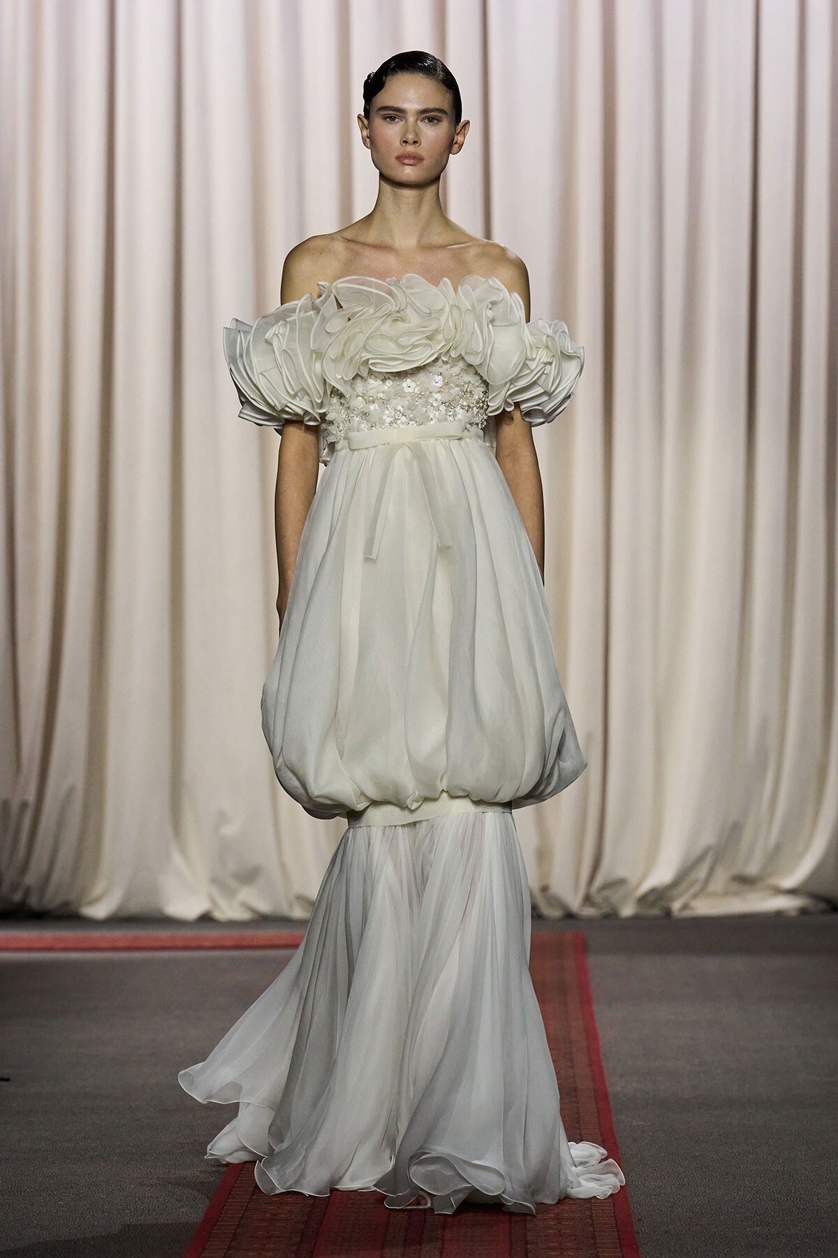 Model on a runway wearing an off-the-shoulder white gown with oversized ruffled sleeves and a voluminous draped skirt.