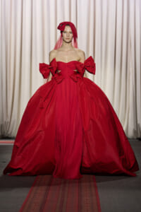 Model on a runway wearing a red ball gown with off-shoulder bodice and oversized bows on the sleeves.