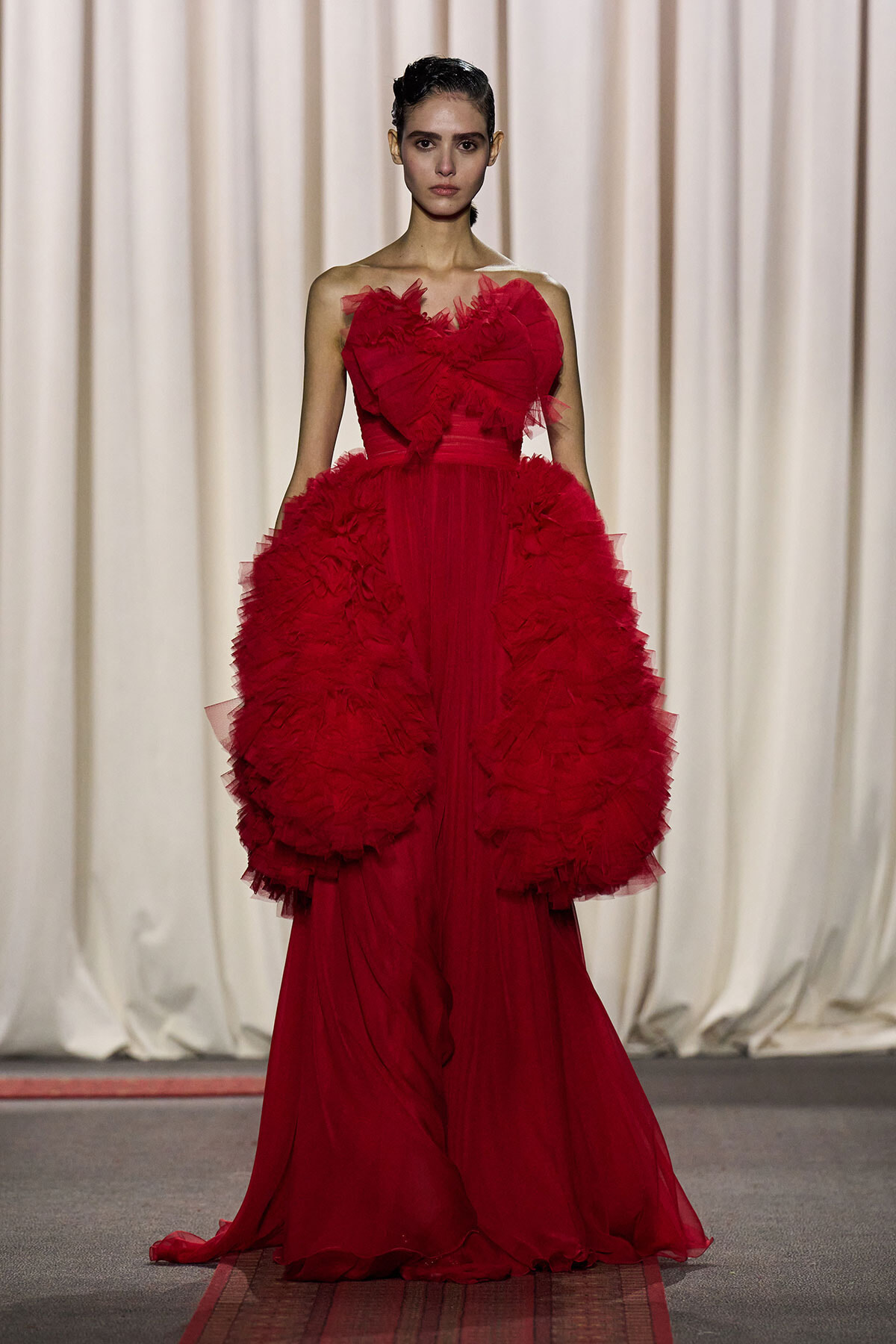 Model walks the runway in a dramatic red ball gown with large ruffled hips and a long train.