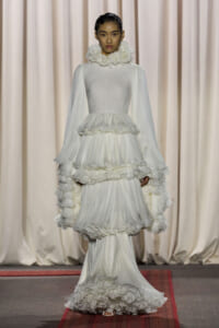 Model walking the runway in a dramatic white gown with tiered ruffles and a high ruffled floral collar.