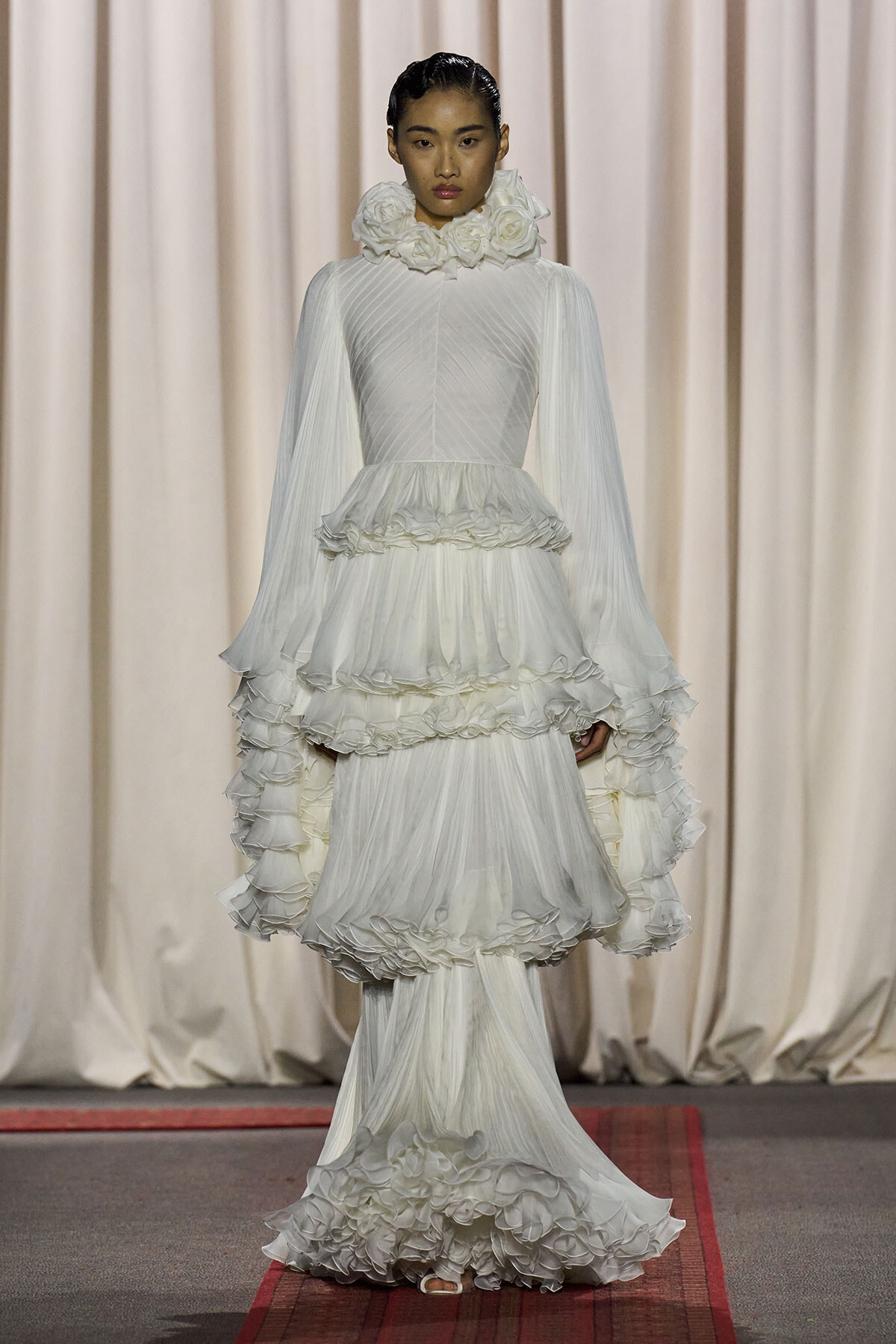 Model walking the runway in a dramatic white gown with tiered ruffles and a high ruffled floral collar.