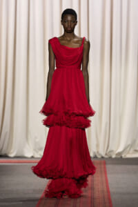 Model on a runway wearing a floor-length red draped gown with multiple layered ruffles cascading down the skirt.