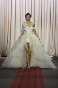 Model walks runway in an off-shoulder white wedding gown with a voluminous, tiered ruffled skirt and veil.