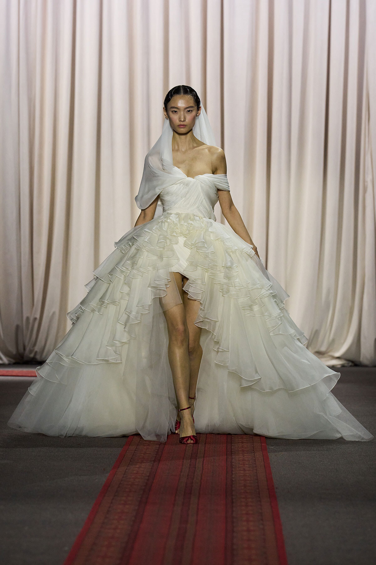 Model walks runway in an off-shoulder white wedding gown with a voluminous, tiered ruffled skirt and veil.