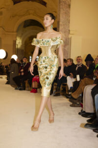 Model walking the runway in a gold off-the-shoulder dress with green floral embroidery, in a grand hall with seated spectators.