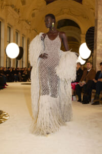 Model walking the runway in a sheer diamond-mesh gown with long white fringe sleeves and hem in a grand ornate hall venue.
