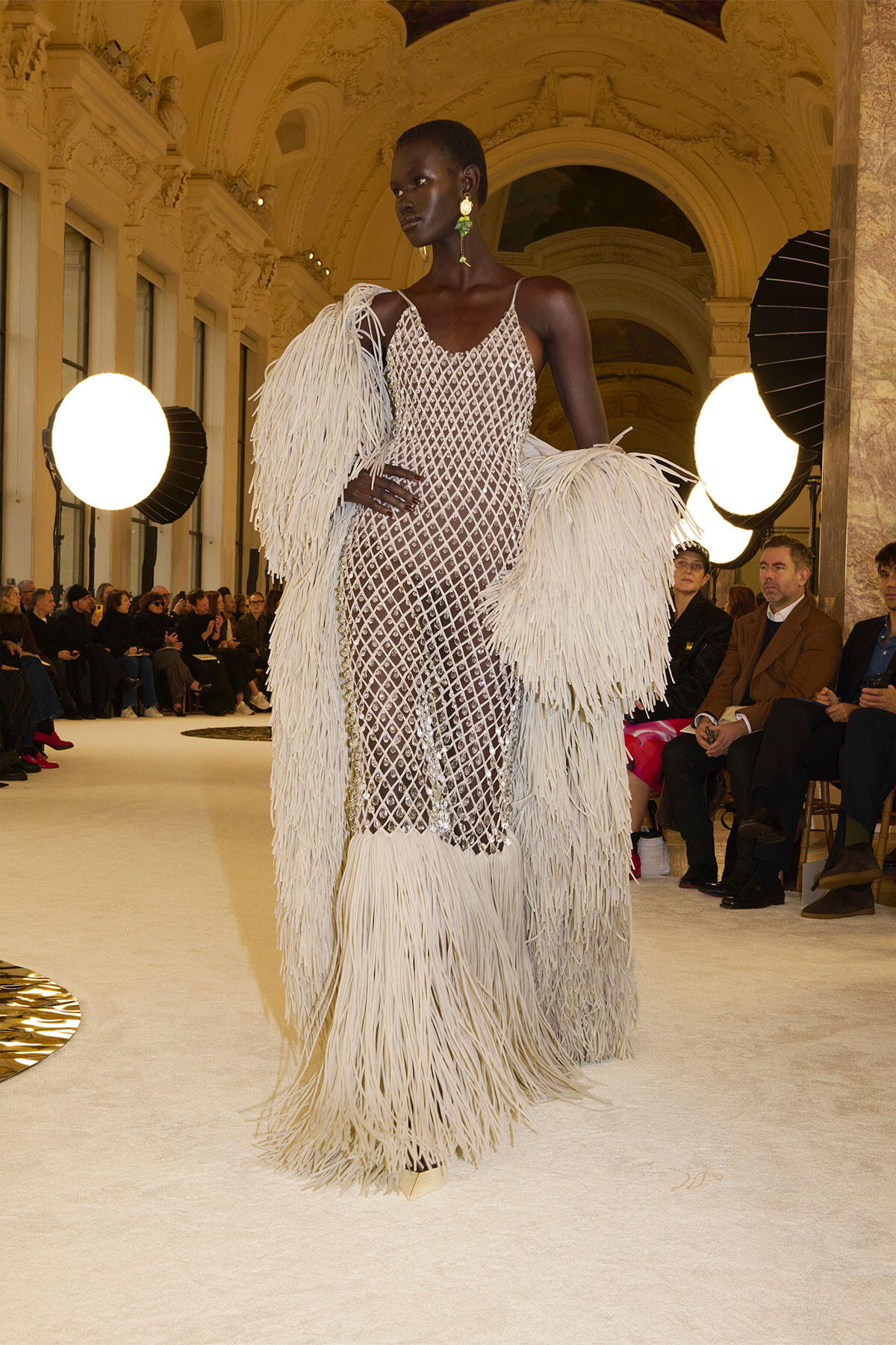 Model walking the runway in a sheer diamond-mesh gown with long white fringe sleeves and hem in a grand ornate hall venue.