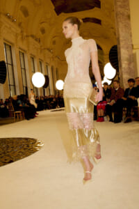 Model walking a runway in a pale, beaded top and tiered gold lace skirt inside a grand hall with round stage lights and an audience nearby.